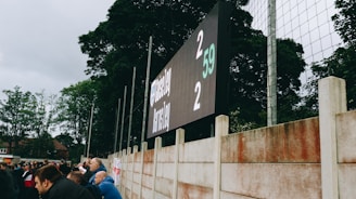 A large digital scoreboard at an outdoor sports venue displays a score of 2-2. Spectators are gathered, observing the event with interest. The scene is enclosed by a tall metal fence and surrounded by lush green trees, indicating a natural setting. Overcast skies add a muted tone to the environment.