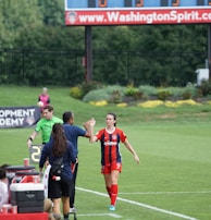 A soccer match scene with a player in a red and blue uniform walking off the field, high-fiving a staff member near the sidelines. A substitution board is held by an official dressed in green. In the background, a scoreboard displays the score, and there is a sign for the Washington Spirit, with greenery and a fence visible further back.