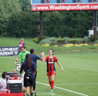 A soccer match scene with a player in a red and blue uniform walking off the field, high-fiving a staff member near the sidelines. A substitution board is held by an official dressed in green. In the background, a scoreboard displays the score, and there is a sign for the Washington Spirit, with greenery and a fence visible further back.