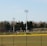 A sports field with a group of people engaging in athletic activities in the background. There is sports equipment like a soccer goal and a scoreboard visible. A chain-link fence runs across the foreground, and a statue or sculpture resembling an eagle is also seen. Trees and tall floodlights are positioned behind the field.