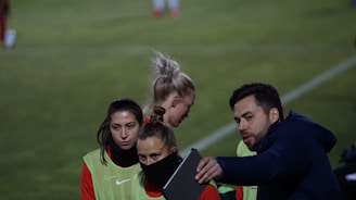A group of individuals on a sports field are wearing green vests over red attire. One person is holding a tablet and seems to be discussing or showing something to the others. The atmosphere suggests a sports event, likely soccer, indicated by their clothing and the setting. The background is a grassy field.