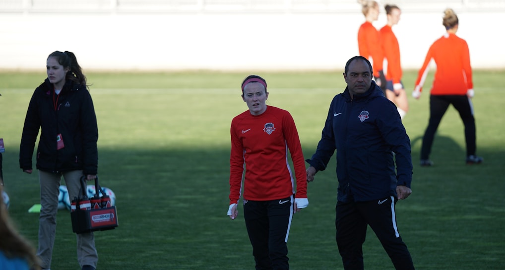 A group of people are gathered on a sports field. Some individuals are dressed in athletic gear, including a woman in a red sports jersey and a man beside her in a blue jacket, appearing to be in conversation. Additional people in similar athletic clothing are visible in the background.