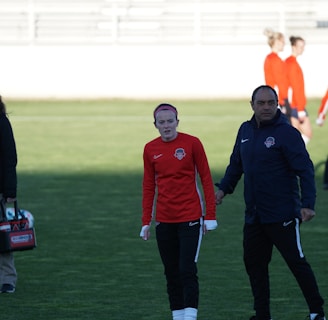 A group of people are gathered on a sports field. Some individuals are dressed in athletic gear, including a woman in a red sports jersey and a man beside her in a blue jacket, appearing to be in conversation. Additional people in similar athletic clothing are visible in the background.