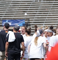 A group of athletes in white shirts with black and red accents are gathered together, engaged in conversation and smiling. They appear to be on a sports field with empty bleachers in the background. The setting is outdoors, and it seems sunny. Some individuals are wearing caps or headbands.