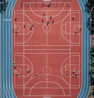 An aerial view of a large, vibrant sports complex featuring a red basketball court with multiple hoops and a surrounding blue running track. Several people are scattered across the area, engaged in various activities.
