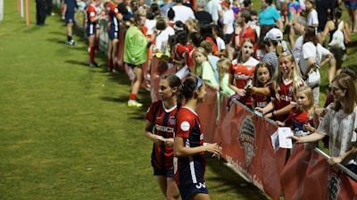The image features a lively gathering of young soccer players and fans next to a field. Several female athletes in red and blue uniforms interact with an enthusiastic crowd standing behind a barrier. Some people in the crowd are holding objects, like pens and paper, possibly for autographs.