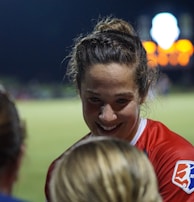 A female athlete in a red sports jersey with the NWSL logo energetically interacts with others on a sports field. The backdrop consists of a darkened night sky and a blurred scoreboard or advertisement display, with soft, colorful lights.