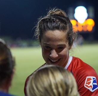 A female athlete in a red sports jersey with the NWSL logo energetically interacts with others on a sports field. The backdrop consists of a darkened night sky and a blurred scoreboard or advertisement display, with soft, colorful lights.