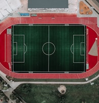 An aerial view of a large sports complex featuring a central green football field surrounded by a red running track. Adjacent to the track, there is a parking area, buildings, and pathways. Nearby, there are additional sports courts in different colors, including a blue swimming pool and orange basketball courts.
