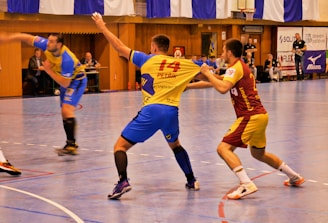 A dynamic indoor sports scene with players in motion on a court. Two athletes in blue and yellow uniforms are engaged in an intense handball game, with one player gripping the other's jersey. The background shows other players, several seated people, and advertisements on the walls.