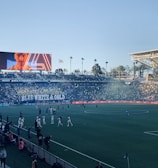A large sports stadium filled with spectators. The field is occupied by soccer players, some walking and others scattered across the pitch. A massive screen displays a person, and flags fly on top of the stadium roof. The audience is dense, and the atmosphere seems lively with a visible banner showing 'BLUE WHITE & GOLD'.