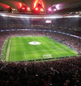 A large, packed stadium with bright lights illuminates a green soccer field. The scene is vibrant with a massive audience surrounding the pitch, visibly excited and engaged. The center of the field displays a prominent circular logo, suggesting a major sporting event. Players and staff can be seen on the field, likely preparing for a match.