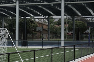 An outdoor sports facility featuring a running track and a soccer goal. The track is red, surrounded by a black metal fence, and the field is covered with artificial green turf. In the background, a basketball court is visible under a covered roof with metal supports. The sky is overcast, adding a moody atmosphere.