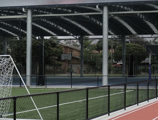An outdoor sports facility featuring a running track and a soccer goal. The track is red, surrounded by a black metal fence, and the field is covered with artificial green turf. In the background, a basketball court is visible under a covered roof with metal supports. The sky is overcast, adding a moody atmosphere.
