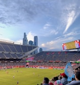 A sports stadium filled with spectators watching a soccer match. Players are on the field in action, and a large scoreboard displays information about the game. The stadium has blue seating and is surrounded by tall city buildings visible in the background against a partly cloudy sky. Fans are dressed casually, some waving flags and wearing team colors.