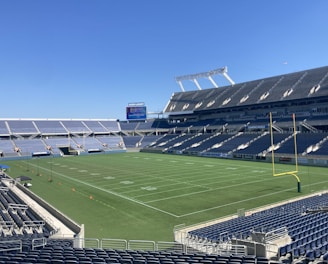 A large, empty sports stadium with blue seats surrounding a well-maintained green grass field. Bright sunlight creates clear shadows, and there are two goal posts on opposite ends of the field. A big electronic scoreboard displays some graphics, and the sky is clear and blue.