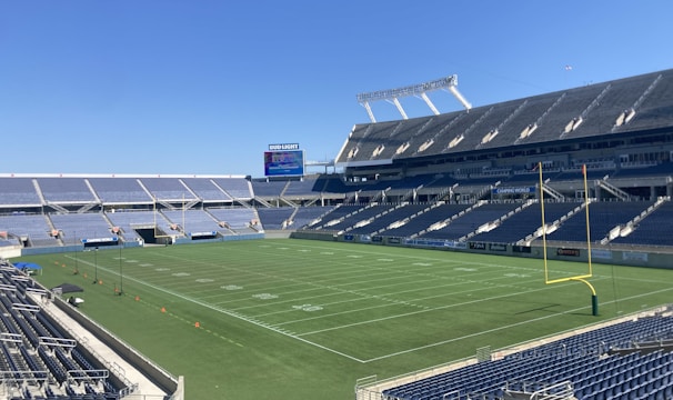 A large, empty sports stadium with blue seats surrounding a well-maintained green grass field. Bright sunlight creates clear shadows, and there are two goal posts on opposite ends of the field. A big electronic scoreboard displays some graphics, and the sky is clear and blue.